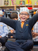 New York Charter Schools - Elementary school scholar with arms out-reached, sitting cross-legged on the floor alongside classmates.
