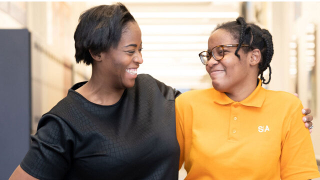 Two people smiling, hugging in a brightly lit hallway.