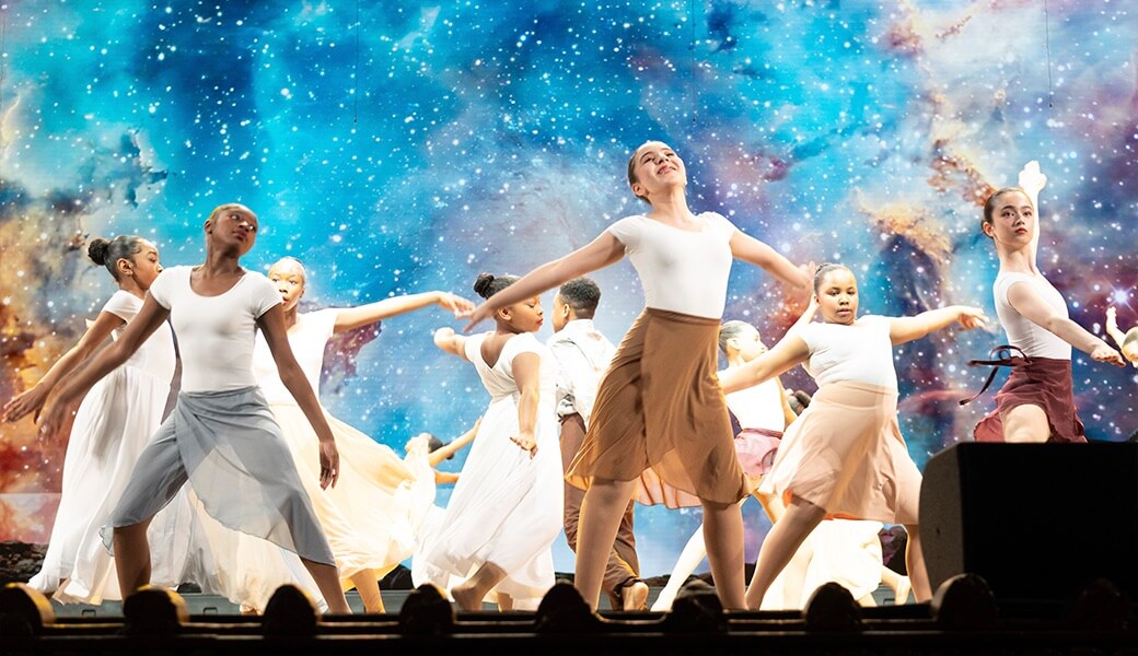 A group of dancers performing a graceful contemporary routine on stage, dressed in flowing skirts and white tops, with a cosmic galaxy backdrop.