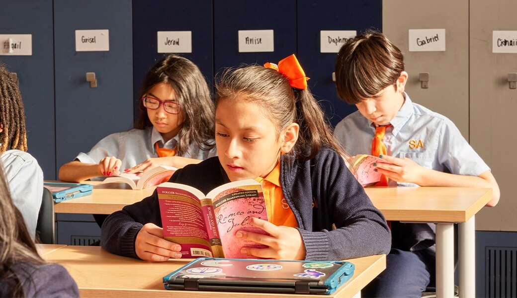 Students in a classroom engaged in independent reading, wearing matching uniforms with orange accents, and seated at desks with books and laptops.