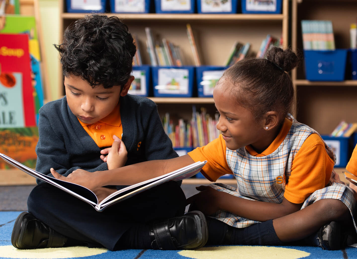 A young boy and girl sit on the floor, reading a book together. The boy, on the left, has dark curly hair and wears a navy blue cardigan over an orange polo shirt. He points to something in the book. The girl, on the right, wears a plaid dress over an orange shirt and looks at the boy as he points.